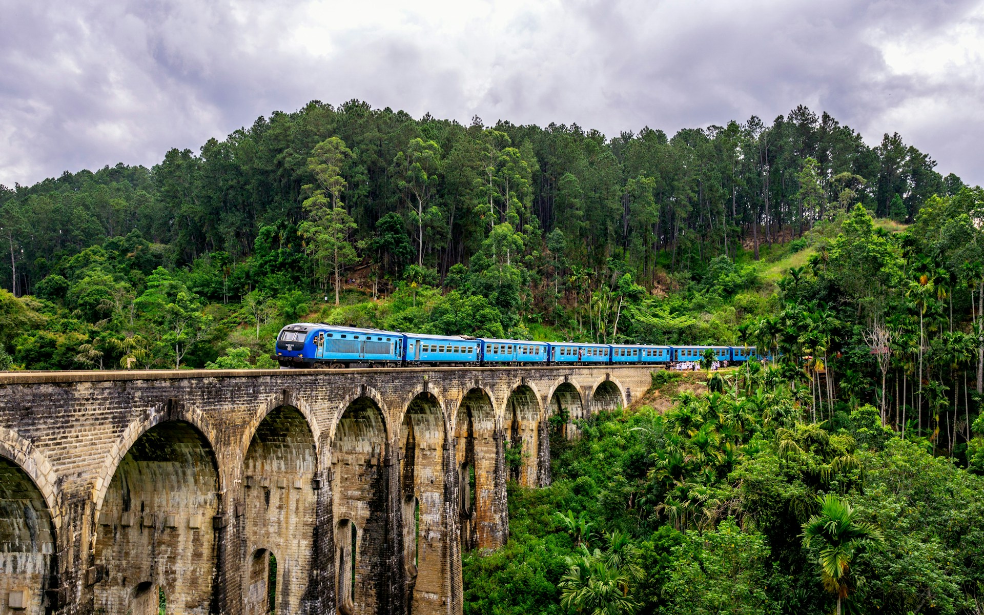 Blauer Zug auf der steinernen Nine Arch Bridge bei Ella, umgeben von dichtem Grün.